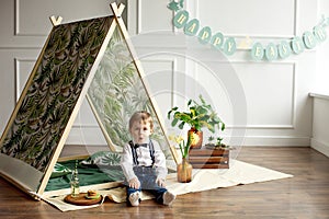 Little boy in a white shirt, jeans and bow tie is sitting in a tent in his room decorated in the style of happy Easter