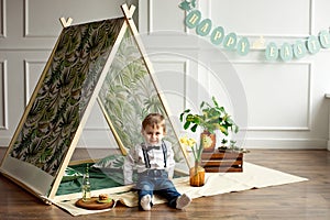 Little boy in a white shirt, jeans and bow tie is sitting in a tent in his room decorated in the style of happy Easter