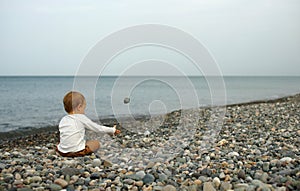Little boy sitting on stone beache and playing with stones. View from behind. Sea and cloudy sky on background