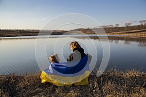 Little boy and a woman are sitting on the shore of a lake, stretching a blue-and-yellow Ukrainian flag