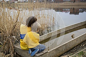 Little boy and a woman are sitting with their backs on the shore of a lake in blue-yellow clothes with a Ukrainian flag