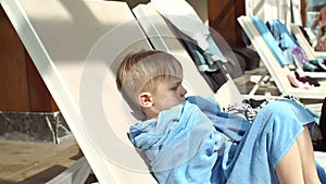 A little boy wrapped in a towel warms up sitting on a lounger by the pool.