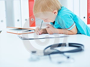 Little boy writes on clipboard sitting at doctor`s desk. Stethoscope in the foreground. Focus on the boy`s hands