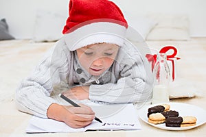 Little boy writing letter for Santa Claus. Child in red santa hat lying on bed and writing christmas letter. Christmas celebration