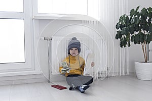 A little boy in a yellow sweater and hat is counting money and studying heating bills, near a heater with a thermostat