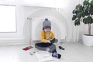 A little boy in a yellow sweater and hat is counting money and studying heating bills, near a heater with a thermostat