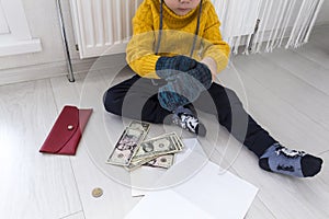 A little boy in a yellow sweater and hat is counting money and studying heating bills, near a heater with a thermostat