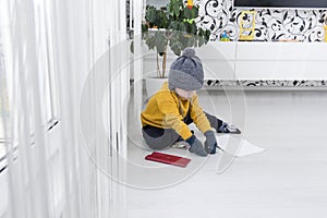 A little boy in a yellow sweater and hat is counting money and studying heating bills, near a heater with a thermostat