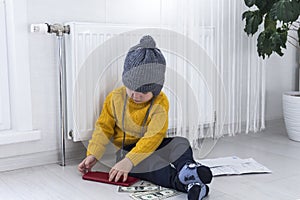 A little boy in a yellow sweater and hat is counting money and studying heating bills, near a heater with a thermostat.