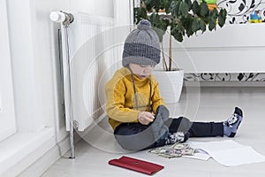 A little boy in a yellow sweater and hat is counting money and studying heating bills, near a heater with a thermostat.