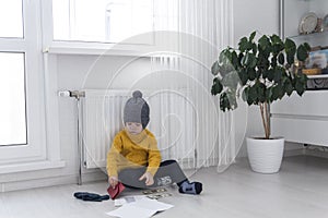 A little boy in a yellow sweater and hat is counting money and studying heating bills, near a heater with a thermostat.