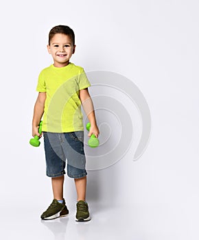 Little boy in yellow t-shirt, denim shorts, khaki sneakers. Smiling, holding green dumbbells behind back, posing isolated on white