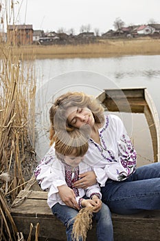 Little boy and young beautiful woman in embroidered national clothes are sitting on shore of lake