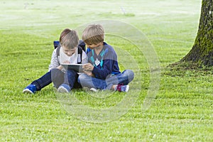 Little boys sitting on the grass in a park and using tablet PC. Technology, lifestyle, education, people concept