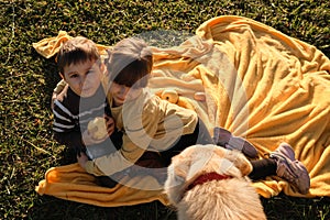 Little brother and sister are sitting in park at sunset on yellow blanket and hugging. Charming Caucasian children. Spend