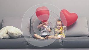 little brothers holding red heart shape balloons playing on grey sofa having fun