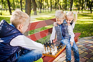 Little brothers playing chess with sister on bench in park. Children intelligence development. Family leisure time. Kids playing