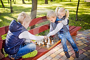 Little brothers playing chess with sister on bench in park. Children intelligence development. Family leisure time. Kids playing