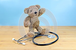 Little brown teddy bear standing on a stethoscope and waving, health care for children, pediatric medicine concept, blue