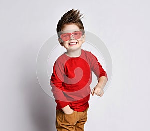 Little brunet boy in sunglasses, red jumper, brown pants. He smiling, hand in pocket, clenched fist, posing isolated on white