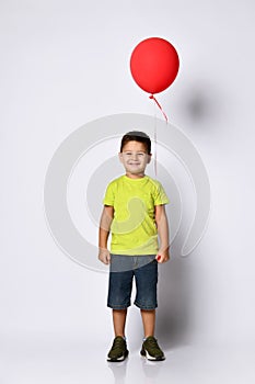 Little brunet boy in yellow t-shirt, denim shorts and khaki sneakers. Smiling, holding red balloon, posing isolated on white