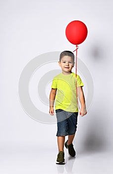 Little brunet boy in yellow t-shirt, denim shorts and khaki sneakers. Smiling, holding red balloon, posing isolated on white