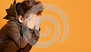 Little brunet child in brown dino hoodie. He has covered his face with palms, posing sitting against orange studio background