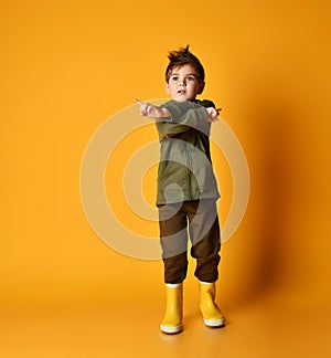 Little brunet child in khaki dino hoodie and pants, yellow rubber boots. He crossed his hands, posing on orange background