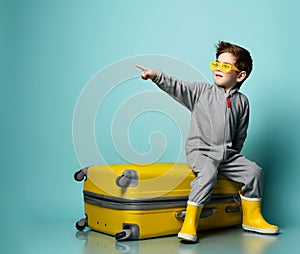 Little brunet kid in gray overall, yellow sunglasses, rubber boots. He sitting on suitcase, pointing, posing on blue background