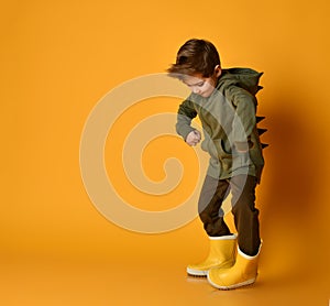 Little brunet kid in khaki dino hoodie with hood and pants, yellow rubber boots. He is posing against orange studio background