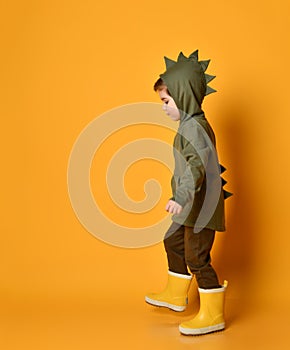 Little brunet kid in khaki dino hoodie with hood and pants, yellow rubber boots. He is walking along orange studio background