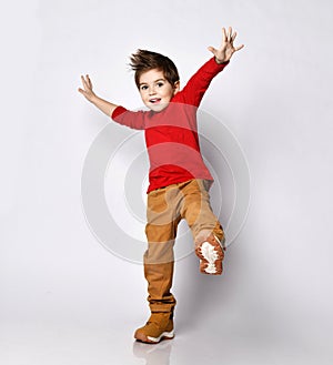 Little brunet kid in red jumper, brown pants and sneakers. He smiling, raised his hands and leg up, posing isolated on white