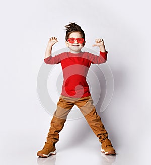 Little brunet kid with stylish hairstyle in sunglasses, red jumper with raised hands up thumbs up opened mouth