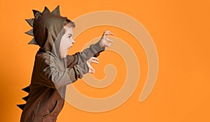 Little brunet male in brown dino hoodie with hood. He growling and scaring you, posing sideways against orange studio background