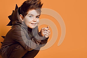 Little brunet male in brown dino hoodie with hood. He is smiling, posing sitting squatting against orange studio background