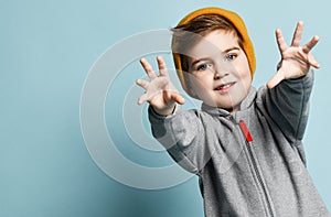 Little brunet male in orange hat and gray overall. He is smiling, reaching his hands to you, posing against blue studio background