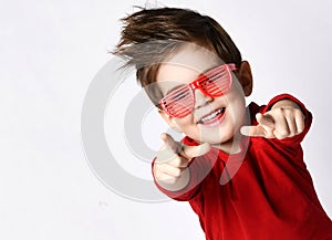 Little brunet male in sunglasses, red jumper. He smiling, pointing at you by forefingers, posing isolated on white photo