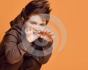 Little brunet model in brown dino hoodie. He growling and scaring you, posing sitting squatting against orange studio background