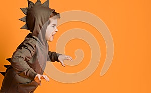 Little brunet model in brown dino hoodie with hood. He roaring and scaring you, posing sideways against orange studio background