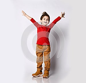 Little brunet model in red jumper, brown pants and sneakers. He smiling, raised his hands up, posing isolated on white background