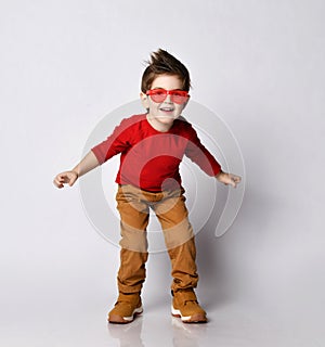 Little brunet model in sunglasses, red jumper, brown pants and sneakers. He smiling, spread hands, posing isolated on white