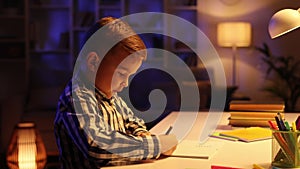 A little brunette boy sits at the table in his room in the evening and draws. A preschooler uses his imagination