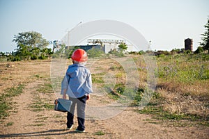 Little builder kid in orange helmet walking towards plant factory industrial with toolbox in hand