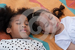 Little Caucasian and African kids girls sleeping and lying together on colorful mat on green grass.