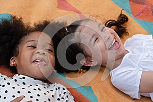 Little Caucasian and African kids girls smiling laughing and lying together on colorful mat on green grass.