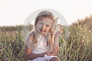 Little caucasian blond girl holding seashell and listening to the sea sitiing on grass field