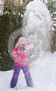 Little Caucasian blond girls in red jacket is playing with snow on fir-tree blur background