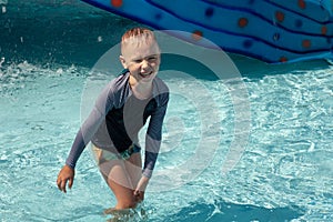 Little Caucasian boy with blond hair laughing in the middle of children pool. Trickles of water everywhere, wet T-shirt and pants, photo