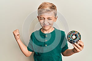 Little caucasian boy kid holding chocolate doughnut screaming proud, celebrating victory and success very excited with raised arm