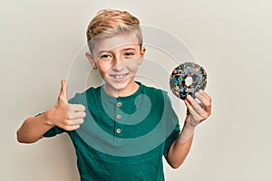 Little caucasian boy kid holding chocolate doughnut smiling happy and positive, thumb up doing excellent and approval sign